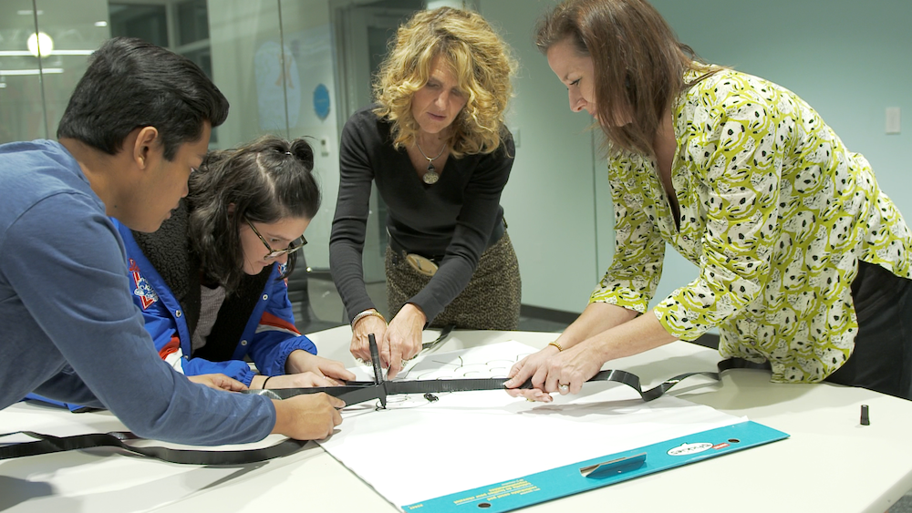Photo of Dr. Leigh Anne Tayler and four students leaning over a oversized pad of paper, using a device made of duct tape and marker to let four of them draw with the same tool at the same time. 