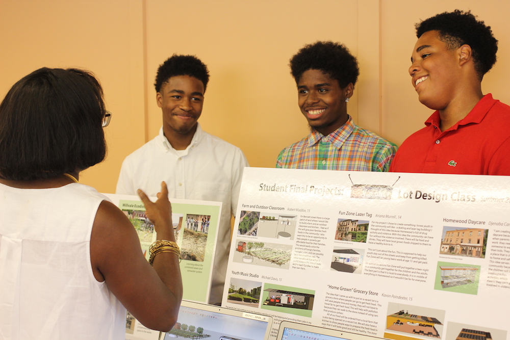 Three students stand proudly behind their research posters and computers describing their work about city lots.
