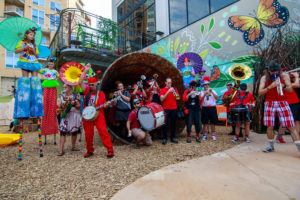A marching band and performers in stilts pose in front of an inviting playground and a nature-inspired mural.