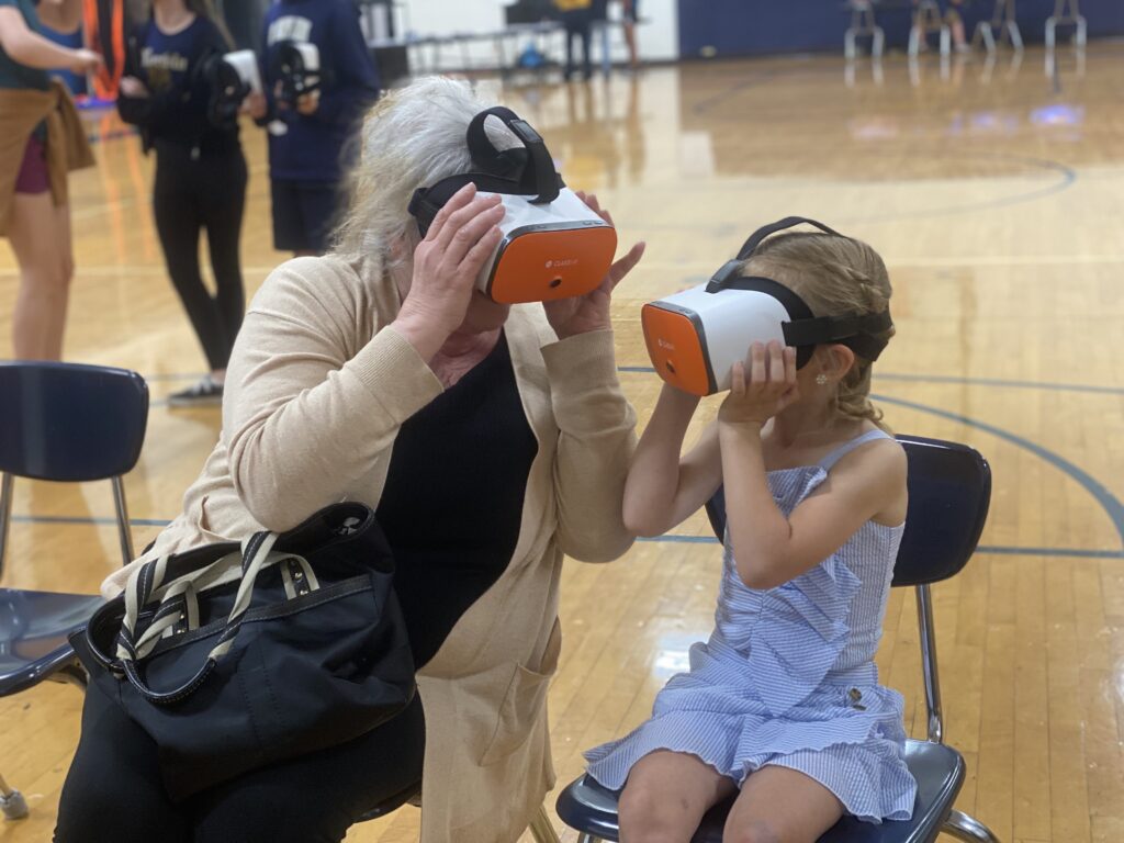 A grandmother and grandchild sit and hold wear VR headsets to their eyes and face each other at a 2022 Norwin event.