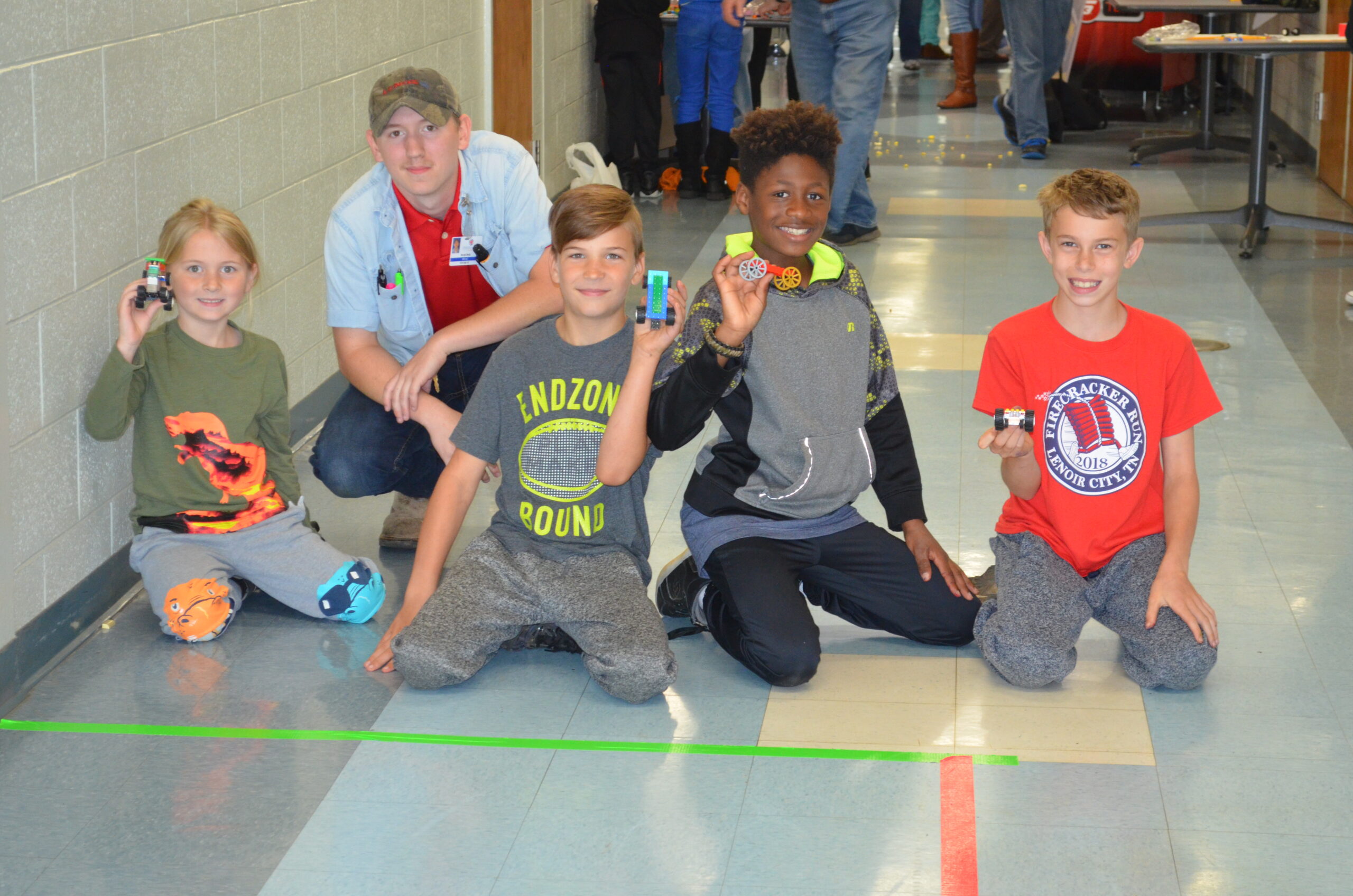 Three children kneel in front of a race course in a school and pose with LEGO cars