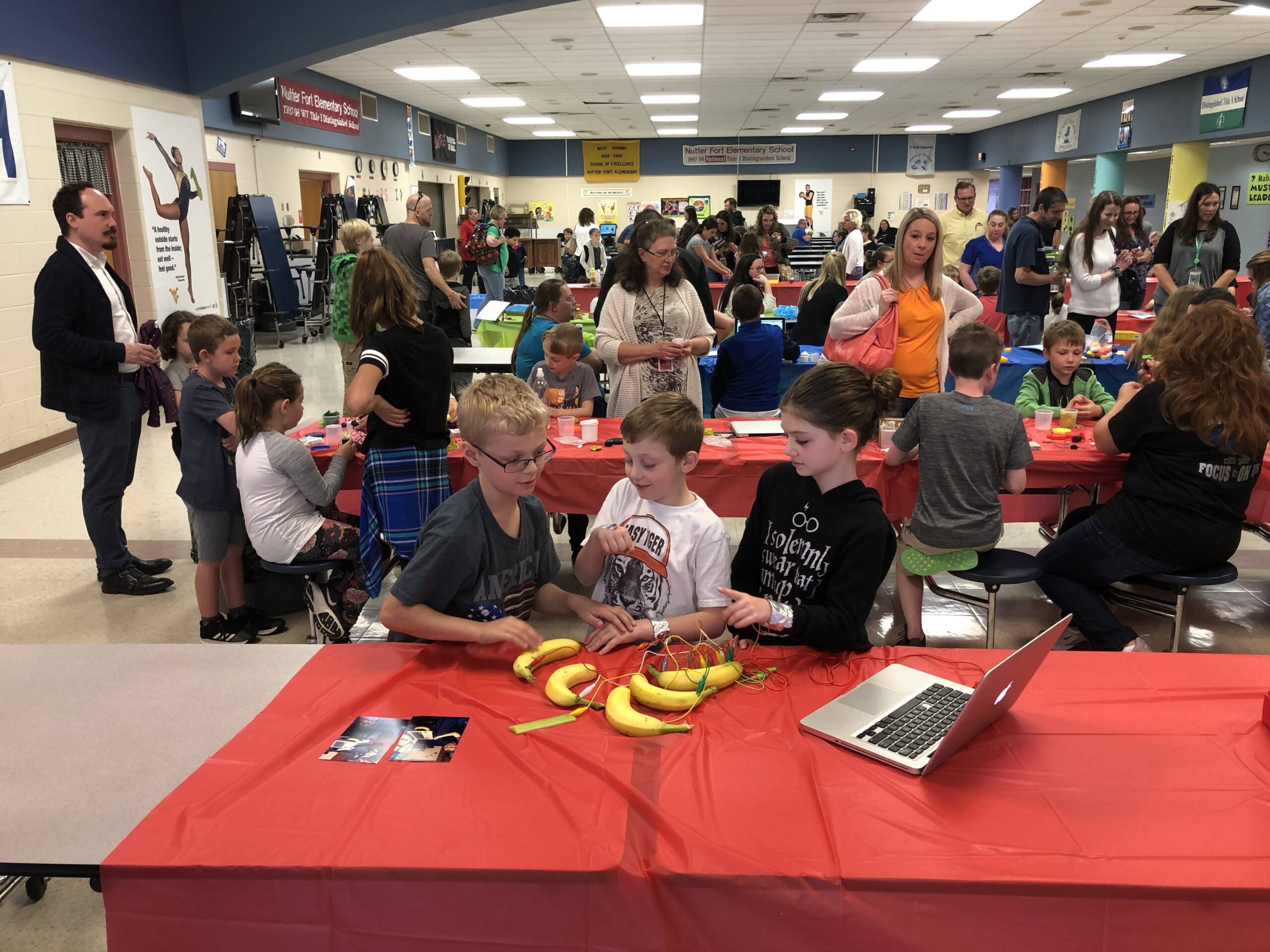 Three children work on a circuit made with wires and bananas. Behind them is a room full of families at work on similar projects.