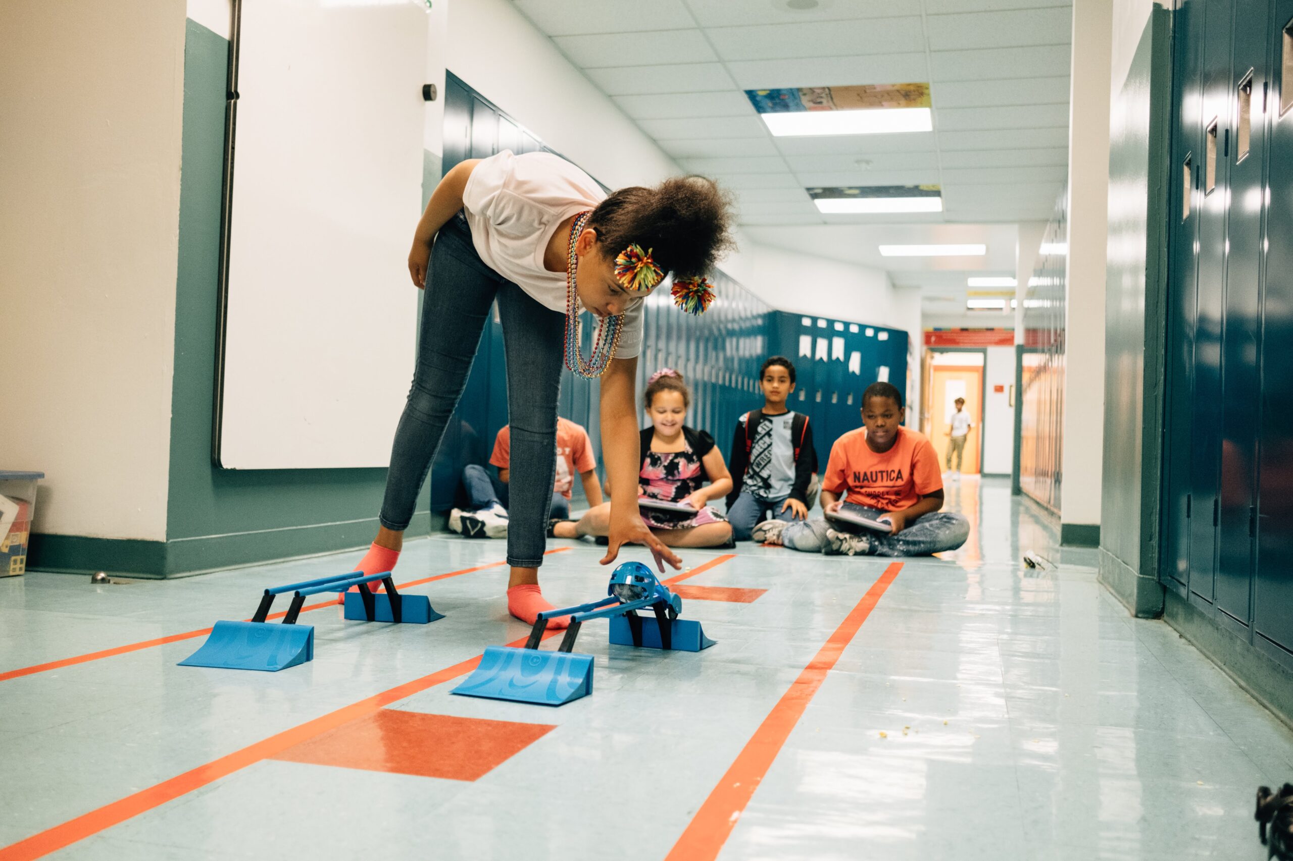 A child bends over to pick up a robot sphere off of a plastic track on the floor. Three children with remote controllers look on in the distance.