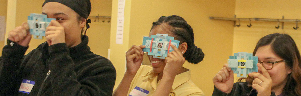 Three participants look through Foldoscopes they hold to their eyes.