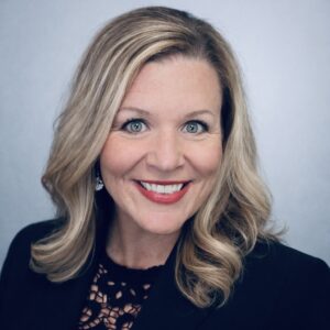 A smiling woman with blonde/brown straight hair just past her shoulders wearing a black blazer