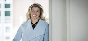 Smiling woman with wavy long dark blonde hair wearing a baby blue backet over a black shirt